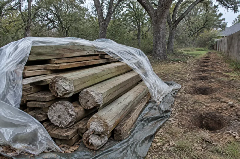 Weathered pressure-treated fence posts with concrete footings stacked on a tarp beside a removed fence line